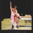 Text: Anna Karenina. Juli | July.

Beschreibung: Ein Tänzer hebt eine Tänzerin, beide in dynamischer Pose auf einer Bühne, nahe einem Tisch mit Glas.