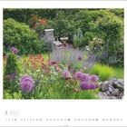 "7 2027": Ein idyllischer Garten mit lila und pinken Blumen, grünem Laub und weißen Gartenstühlen im Hintergrund.