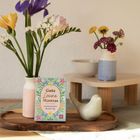 "Gute Laune Mantras - Sonnenschein für jeden Tag" steht auf einer Karte. Blumen in Vasen und eine Taubenfigur umgeben sie.