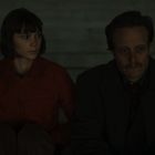 Zwei Personen sitzen nebeneinander auf Stufen. Links eine Frau in roter Jacke, rechts ein Mann im Mantel mit nachdenklichem Ausdruck.