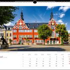 Juli-Kalenderblatt mit einem roten Gebäude auf einem Platz, blauer Himmel, Bäume und eine Statue im Vordergrund.