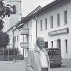 "Unser Gasthaus - noch immer steht das Gebäude" steht neben einem Kirchturm und einer Straße mit parkendem Auto und Mann.