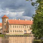 Ein Schloss mit rotem Dach spiegelt sich in einem ruhigen See, flankiert von einem Baum. Der Himmel ist bewölkt.