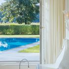 Ein Raum mit offenem Fenster zeigt einen Pool, dahinter ein Baum und Hecke. Innen sind Vorhänge und ein Mosaiksitz mit Becken.