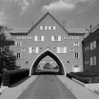 Gebäude mit Spitzbogen, symmetrischer Bauweise, Fenstern mit Fensterläden. Gepflegte Hecken und befahrbare Straße darunter.