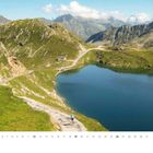 Text "Juli", Kalenderzahlen unten. Berglandschaft mit See, Radfahrer auf einem Pfad, grasbewachsene Hügel, klare Himmel.