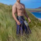 "Juli" steht unten. Ein muskulöser Mann ohne Hemd trägt einen Kilt in einer grünen Landschaft mit blauem Himmel.