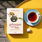 Buchcover: "Geborgen und frei" von Pierre Stutz. Daneben eine Tasse Tee auf einem gelben Tisch.