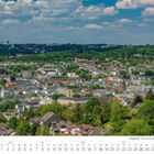 Kalender zeigt Juli 2027. Blick über Stadt mit vielen Häusern, dahinter grüne Hügel und blauer Himmel mit Wolken.