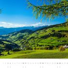 "Blick über das Villnößtal zum Eisacktal und auf die Sarntaler Alpen." Ein grünes Tal mit verstreuten Häusern und Bergen im Hintergrund.