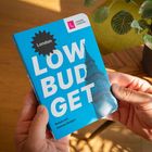 Auf einem blauen Buch stehen "London LOW BUDGET", "Reisen mit kleinem Budget". Eine Hand hält es, daneben eine Pflanze.