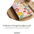 Exklusives Design handgemacht. Notizbuch mit Blumenmotiv, grünem Gummiband, Holzuntergrund, Stift daneben.