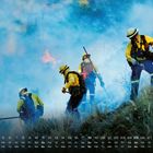 Kalender mit Datum. Feuerwehrleute in gelber Schutzkleidung bekämpfen Flammen und Rauch in einer grasbewachsenen Landschaft.