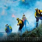 Kalender mit Daten und Wochentagen unten. Feuerwehrleute in gelber Kleidung bekämpfen Flammen und Rauch im Wald.
