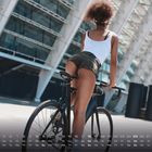 Eine Frau in Shorts und einem weißen Top fährt auf einem Fahrrad. Hintergrund: modernes Gebäude, unten Kalenderdaten.