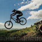 Kalenderdaten unten, Tag „So“ hervorgehoben. Ein Mountainbiker springt von einer Rampe über eine grüne Landschaft.