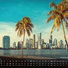 Kalender: Skyline Miami Beach im August, moderne Gebäude hinter Palmen am Wasser, blauer Himmel.