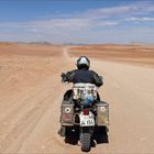 Nummernschild: "WM A 114". Ein Motorradfahrer fährt auf einer einsamen, staubigen Straße durch eine weite Wüstenlandschaft.