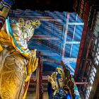 Eine große, goldene Buddhastatue mit gefalteten Händen, verziert mit bunten Tüchern und Perlen, in einem farbenprächtigen Tempel.