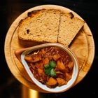 Ein ovaler Teller mit Gulasch, garniert mit Petersilie. Zwei Scheiben Brot liegen auf einem runden Holzbrett.