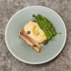 Ein kunstvoll angerichtetes Gericht: Creme, Fischfilet, Kapern und grüne Blätter auf einem grauen Teller.