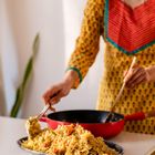 Eine Person in bunter Kleidung serviert Reis aus einer roten Pfanne auf einen Teller.