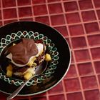 Ein Teller mit Schokoladenkuchen, Sahne und Mangostücken, auf rotem, gekacheltem Tisch, mit einem Löffel daneben.