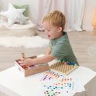 Ein Kind spielt an einem Tisch mit einer Montessori-Holzplatte und farbigen Murmelvorlagen in einem hell eingerichteten Zimmer.