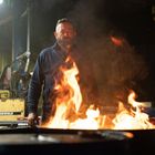 Im Hintergrund: "SUREWELD" auf gelbem Kasten. Ein Mann steht hinter einem Fass mit loderndem Feuer. Industrielle Umgebung.