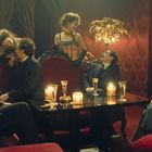 Keine sichtbaren Texte. Eine Gruppe elegant gekleideter Menschen in einem luxuriösen Raum mit rotem Samt und Kerzenlicht.