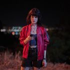 Eine Frau hält ein Handy, trägt eine rote Jacke mit "Eldborg Academy". Hintergrund: verschwommene Lichter einer Stadt.