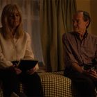 Eine Frau und ein Mann sitzen auf einem Bett in einem abgedunkelten Raum, die Frau hält ein Buch. Ein Lampenlicht erhellt die Szene.