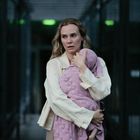 Eine Frau in heller Kleidung hält ein Baby in rosa Decke in einem dunklen Flur. Sie wirkt besorgt oder nachdenklich.