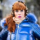 Eine Frau mit roten Haaren trägt einen blauen, glänzenden Daunenmantel und Leoparden-Ohrenwärmer.