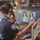 Am Computer zeigt eine Polizeibeamtin zwei Frauenbilder und telefoniert. Der Schreibtisch ist mit Büromaterial gefüllt.