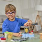 Ein Junge mit blauer Brille und blauem T-Shirt spielt fröhlich mit Knete an einem Tisch in einem gemütlichen Wohnzimmer.