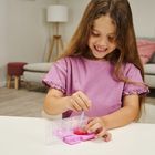 Ein lachendes Mädchen in einem lila Shirt rührt mit einem Stäbchen in einem rosa Behälter am Tisch.