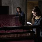 Eine Frau an einem Webstuhl mit roten Fäden, ein Mann im Hintergrund; beide in antik inspirierter Kleidung.