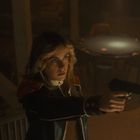 Eine Person mit langen Haaren zielt mit einer Pistole. Im Hintergrund ein Tisch mit Stühlen, schwach beleuchtet.