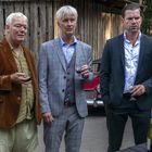 Drei Männer in Anzügen halten Weingläser. Sie stehen draußen in der Nähe eines Tisches mit Weinflaschen.