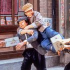 Zu sehen sind chinesische Schriftzeichen an einem Gebäude. Drei Männer sind in einer dynamischen, kämpfenden Szene.