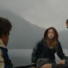 Drei Personen in Regenkleidung sitzen in einem Boot auf einem nebligen See, umgeben von Bergen. Sie wirken angespannt.