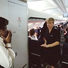 Das Bild zeigt den Innenraum eines Flugzeugs. "NO SMOKING IN LAVATORY" auf einer Tür. Eine Flugbegleiterin spricht mit einer Frau. Passagiere sitzen.