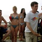 Ein junger Mann trägt ein weißes Shirt mit einem bunten Drachenmotiv und gestikuliert; um ihn stehen mehrere Jugendliche am Strand.