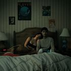 An der Wand diverse Poster, eins mit "PLAYER". Ein Paar sitzt auf einem Bett, sie lehnt sich an ihn. Dämmerbeleuchtung.