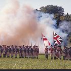 Soldaten in historischen Uniformen stehen in Formation auf grünem Feld, hinter ihnen steigt Rauch auf, Union Jack weht neben ihnen.