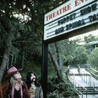 "Theatre Entrance," "Puppet Show and Spinal Tap" auf einem Schild. Drei Personen blicken nach oben im Freien.