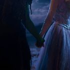 Zwei Personen stehen Hand in Hand; die eine trägt ein schwarzes, die andere ein funkelndes Abendkleid. Es ist Nacht.