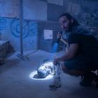 Ein Mann kniet mit einer Taschenlampe neben einer Mumie in einem blauen, staubigen Raum mit mysteriösen Zeichnungen an der Wand.