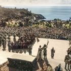 In einer mittelalterlichen Stadt mit Flaggen versammeln sich viele Menschen und Ritter auf einem Platz. Im Hintergrund das Meer.